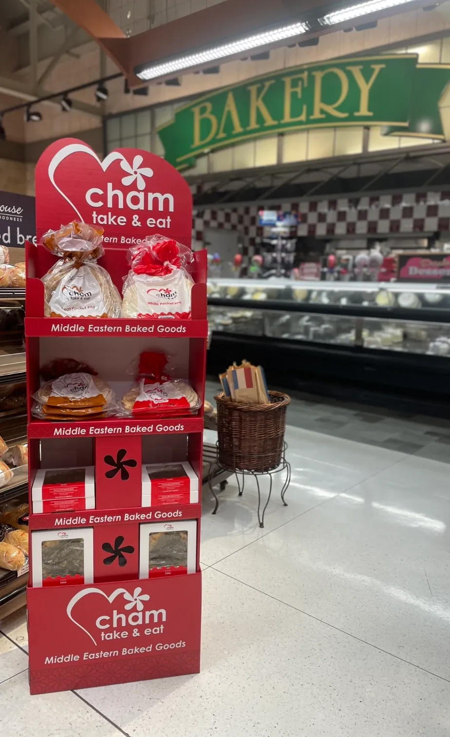 A red Cham Bakery display stand in a grocery store's bakery department, its shelves stocked with bags of white and whole wheat pocket pita and boxes of za'atar bread