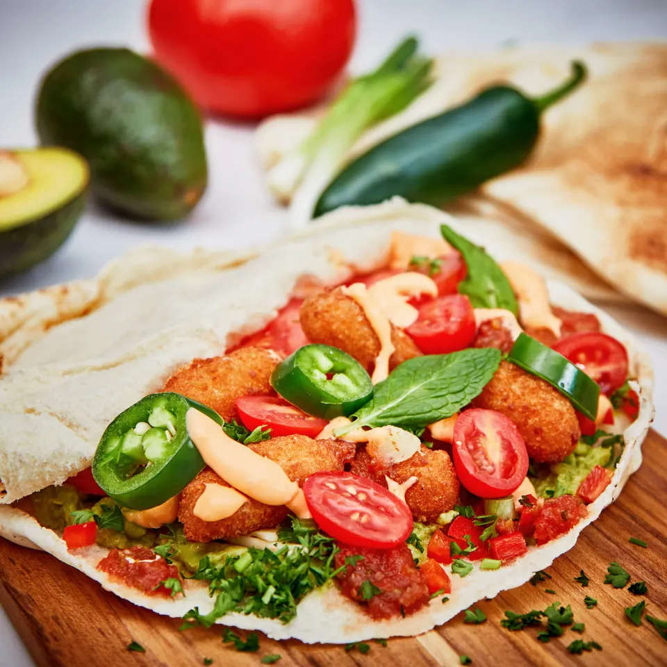 A pita topped with falafel, peppers and sauce, with avocado and tomato behind